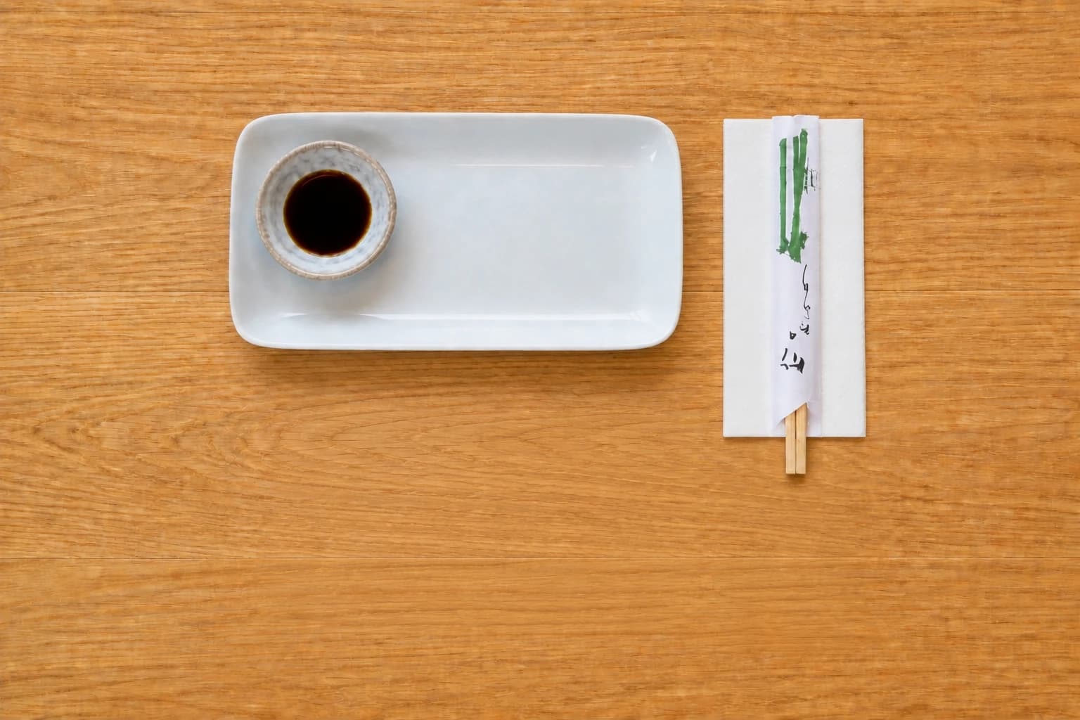A place setting near the edge of a wooden table: Enso's rectangular plate with one soy sauce dish on the left, and the restaurant's paper-wrapped bamboo chopsticks on a napkin on the right.